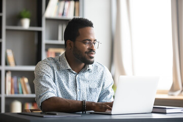 Satisfied African American man wearing glasses looking at computer screen, reading good news in...