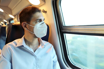 Fototapeta na wymiar Travel safely on public transport. Young man with KN95 FFP2 face mask looking through train window. Train passenger with protective mask looking through the window.