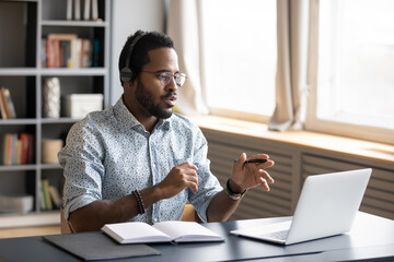 African American man wearing headphones speaking, using laptop, student wearing glasses learning language, watching video webinar or listening to lecture, mentor coach holding online lesson