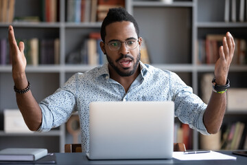 Irritated angry African American man wearing glasses having problem with broken laptop, confused...