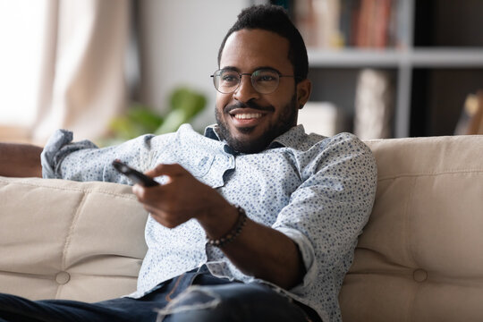 Smiling African American man wearing glasses holding using tv controller, sitting on cozy couch in modern living room, watching movie or football match at home, spending leisure time with television