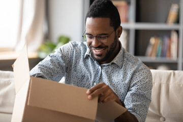 Smiling African American man wearing glasses unpacking parcel, sitting on couch at home, happy...