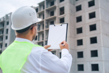 Chief in a vest and a construction helmet with documents in his hands, against the background of a tower crane and the sky with clouds. House construction inspection.