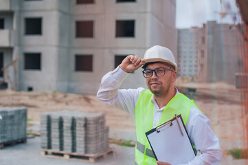 Chief in a vest and a construction helmet with documents in his hands, against the background of a tower crane and the sky with clouds. House construction inspection.