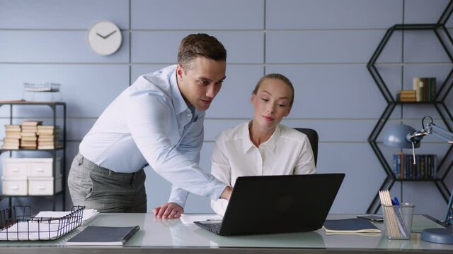 young man and woman work together in office with laptop on desk. he points to screen, she looks and nods. looks positive and cheerful, smiles. look to camera. discuss and brainstorm   