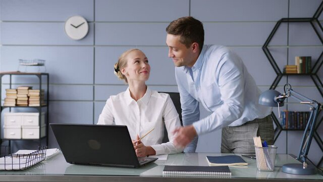young man and woman work together in office with laptop on desk. he shows thumb up, points to screen, she looks and nods. looks positive and cheerful, smiles. discuss and brainstorm 