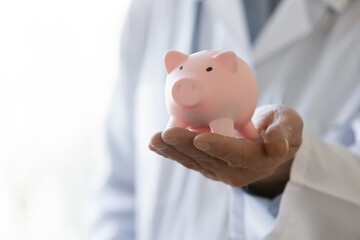 Close up mature doctor physician gp wearing white coat uniform holding pink piggy bank, medical insurance, healthcare money savings concept, finance, hospital budget and accounting