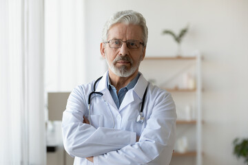 Head shot portrait mature doctor wearing glasses standing with arms crossed in office, confident...