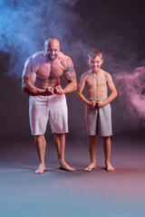 Powerful muscular man shirtless posing with his son in the studio with red and blue smoke