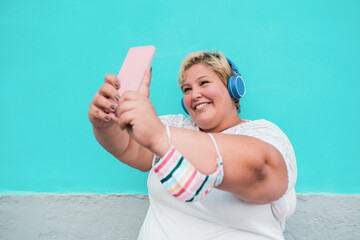 Young curvy woman taking a selfie with smartphone while wearing headphone - Beautiful plus size girl laughing