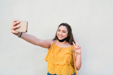 Cheerful asian woman laughing and taking a selfie with smartphone while doing the peace symbol with hand  - Happy influencer girl wearing protective face mask