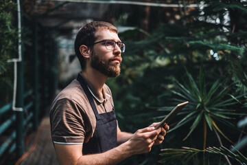 Young man gardener in glasses and apron with digital tablet working in a garden center for better quality control. Environmentalist using digital tablet in greenhouse.