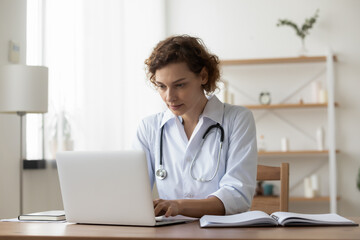 Serious woman wearing white uniform with stethoscope working on laptop, sitting at desk in...