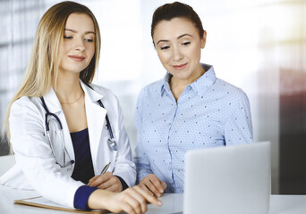 Obraz premium Young beautiful woman-doctor and her patient are discussing patient's current health examination, while sitting together at the desk in the sunny cabinet in a clinic. Female physician is writing some