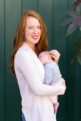 Medium vertical shot of pretty smiling red-haired young woman holding her newborn baby girl tenderly while standing in front of dark green wall