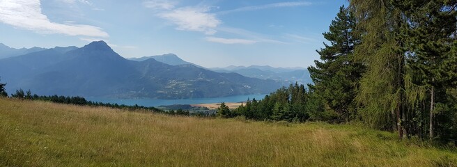 Panoramique : champs devant le lac, la lisière de la forêt et les montagnes