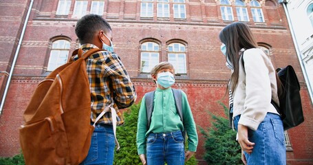 Portrait of multi-ethnic children in medical masks greeting with elbows near school in quarantine. Cute African American and Caucasian boys outdoors. Beautiful girl with friends. School concept