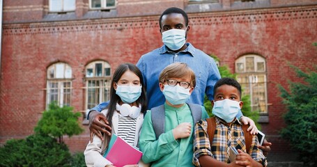 Portrait of mixed-races junior students standing with male teacher near school in masks in...