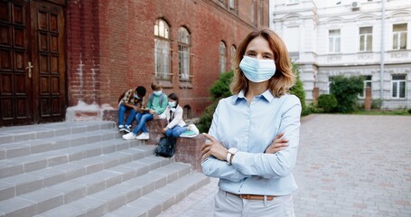 Portrait of beautiful Caucasian happy female teacher in mask smiling to camera in front of school. Woman standing outdoor at schoolyard. Mixed-races junior students on background. Pandemic concept