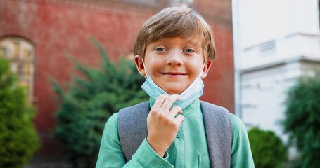 Close up of cheerful Caucasian cute boy putting off medical mask while standing near school and...