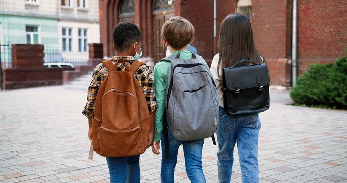 Rear of multi-ethnic junior students with backpacks going to school. Caucasian girl and boy. African American pupil with schoolbag. Male and female teachers on background tapping on smartphone