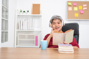 Fototapeta na wymiar Relax elderly Asian man reading a book and listening to music in home office.