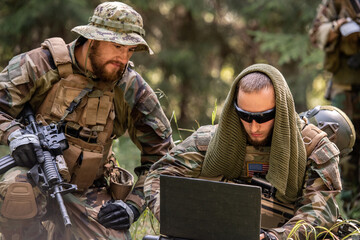 Army American soldiers in camouflage outfits sitting on ground and using laptop while discussing...