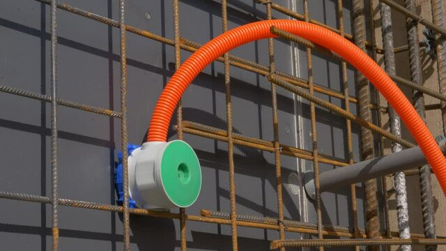 CLOSE UP: Thin corrugated orange conduit runs out of an electricity outlet installed on the outer side of an unfinished wall of a house under construction. Electricity conduit and rusty metal wiring.