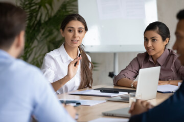 Confident young arab ethnicity woman sitting at table with diverse colleagues, explaining marketing strategy or developing project ideas at brainstorming meeting, office communication concept.