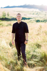 Full length photo of handsome young man in black shirt and pants, standing outdoor in the beautiful green summer field at sunset