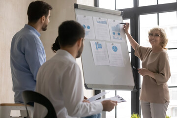 Smiling happy middle aged old female leader coach pointing at marketing research graphs on...