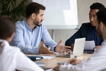 Skilled handsome 30s businessman talking to multiracial partners in boardroom. Young diverse mixed race employees sharing ideas at brainstorming meeting, sitting together at table in office.