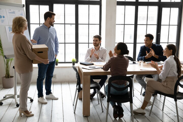Smiling young male leader introducing middle aged female new worker holding box with stuff to group of international colleagues at meeting in modern office, acquaintance or retirement concept.