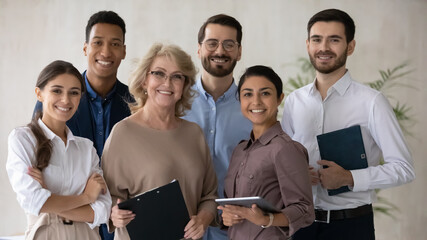 Portrait of smiling 60s middle aged mature female mentor standing in office with motivated skilled young mixed race staff of international company, happy diverse business people looking at camera.