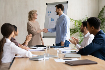 Happy young mixed race teammates sitting at table, applauding while watching smiling male leader shaking hands with older 60s female colleague or partner, celebrating good deal or career promotion.