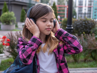 small / young beautiful brunette girl in a plaid pink shirt closed her eyes and listens to music with headphones in the summer outdoors in the park