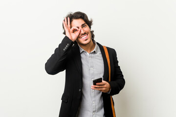 Young business man holding a phone excited keeping ok gesture on eye.