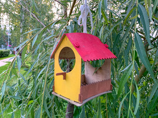A small birdhouse with a red roof and yellow walls hangs on a willow branch. Caring for nature, food for birds.