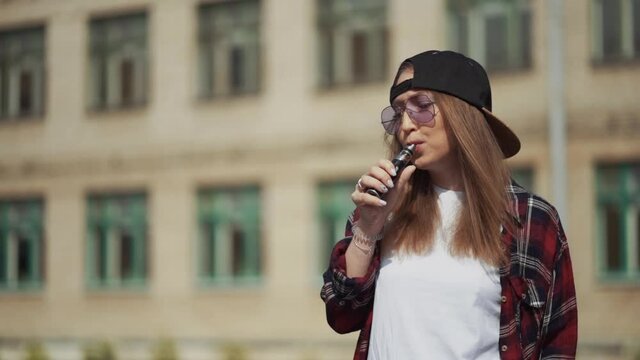 Pretty young hipster woman in black cap vape ecig. Girl vape popular ecig gadget, vaping device.Happy brunette vaper girl with e-cig. Portrait of smoker female model with electronic cigarette