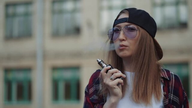 Close up pretty young hipster woman in black cap vape ecig. Girl vape popular ecig gadget, vaping device.Happy brunette vaper girl with e-cig. Portrait of smoker female model with electronic cigarette