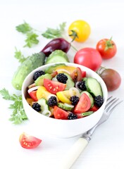 Vegetable berry salad of fresh yellow, red, black tomatoes, cucumbers, onions and blackberries in a bowl on a white background