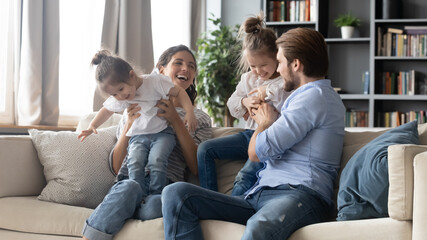 Joyful carefree parents playing with adorable little kids, resting on comfortable couch together at...
