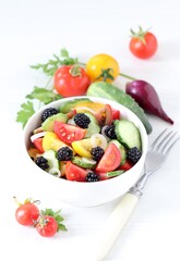 Vegetable berry salad of fresh yellow, red, black tomatoes, cucumbers, onions and blackberries in a bowl on a white background