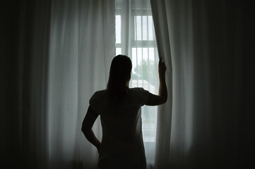 Fototapeta na wymiar Caucasian woman at the window in semi-darkness in the morning opens the curtains.