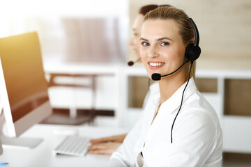 Blond female customer service representative is consulting clients online using headset in sunny office. Call center concept