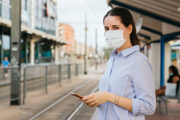 Fototapeta na wymiar Stock photo of a young woman wearing a face mask, holding a phone and waiting on a tram station