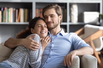 Loving devoted young handsome man cuddling attractive happy curly woman, enjoying lovely sweet moment together indoors. Smiling relaxed family couple spending leisure time at home, relations concept.