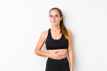 Young caucasian fitness woman posing in a white background touches tummy, smiles gently, eating and satisfaction concept.