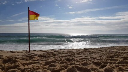 Clear sand beach with yellow and red caution flags flapping in the wind. The waves crash on the shore and the sun reflects off the water. 