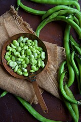 Small copper pan on hessian cloth on a brown background.  Green broad beans pods enter the frame.